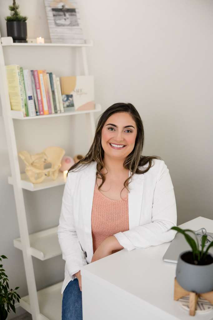 Bruna Gomes smiling at her desk, beside a shelf of books and a pelvis model